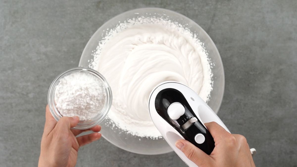 glass bowl of whipped cream with mixer and bowl of powdered sugar held over top