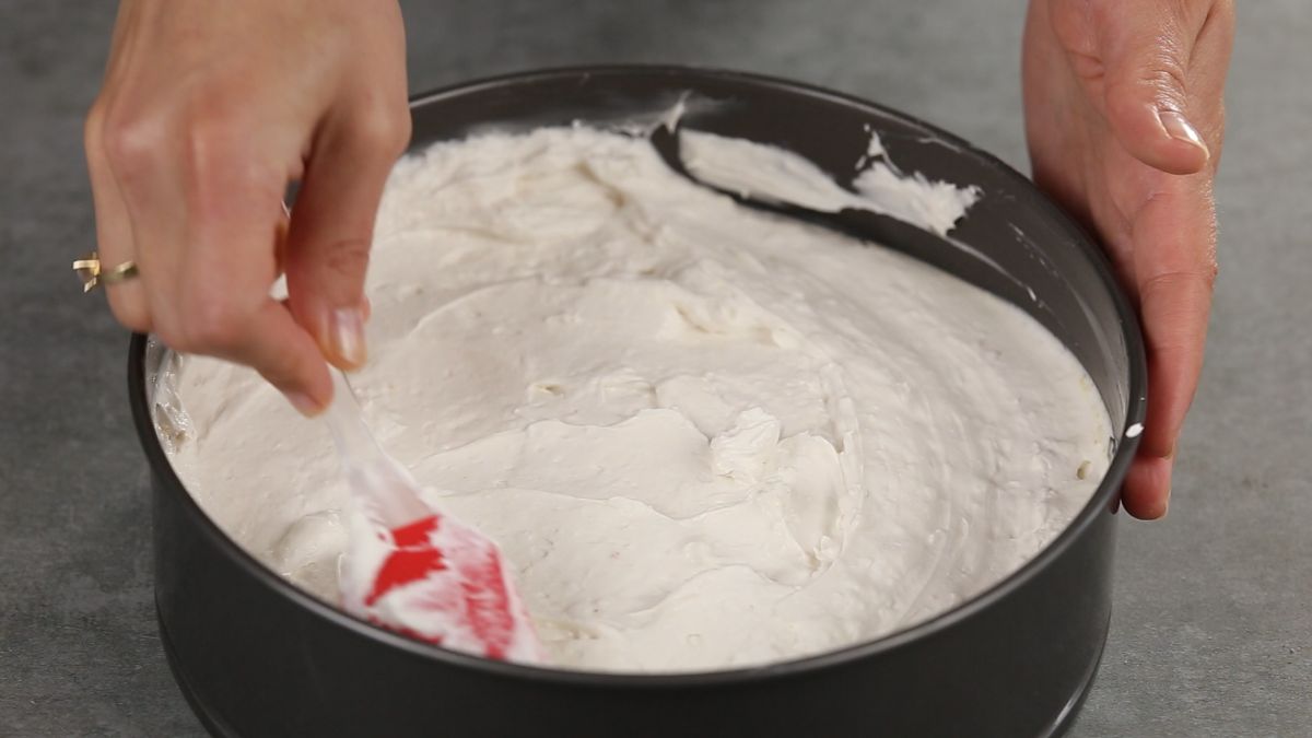 hands using red spatula to spread cheesecake over crust in springform pan