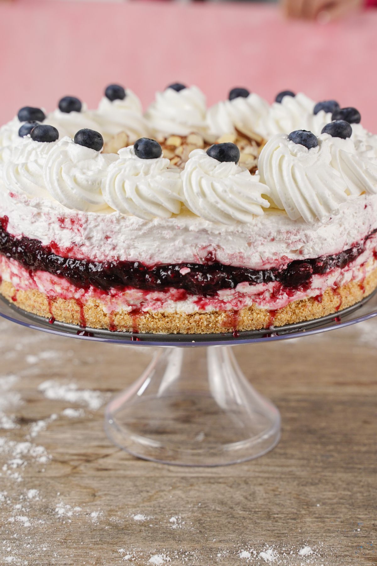 blueberry cheesecake topped with whipped cream, almonds, and berries on a glass cake platter