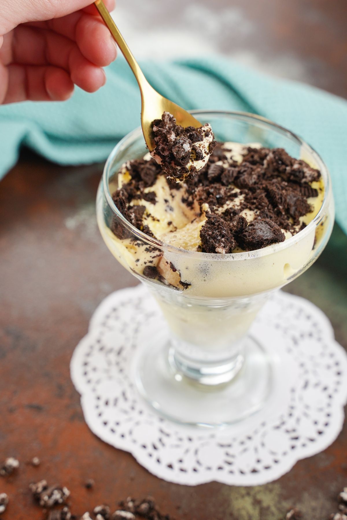 dessert glass of pudding and Oreo cookies on white doily with spoon held over the top