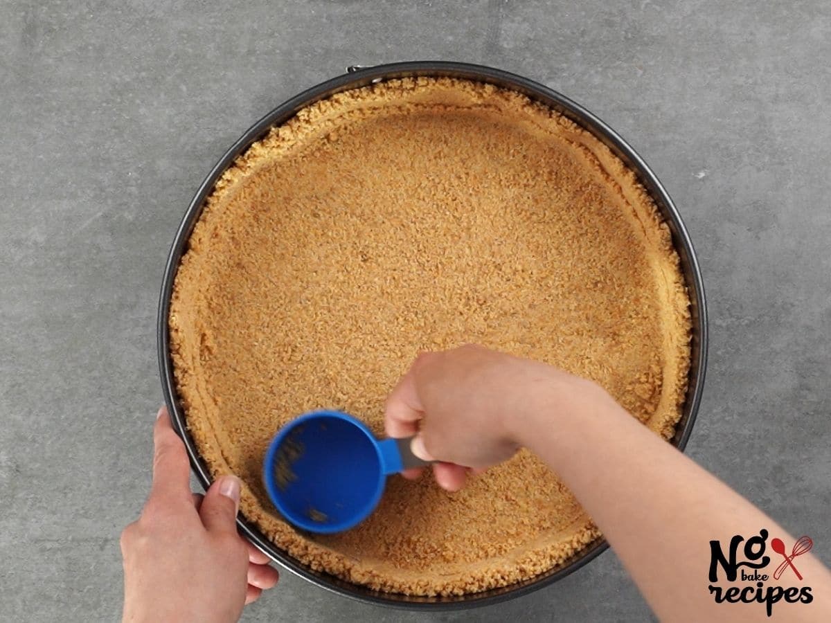 blue measuring cup pressing cookie crumbs into pie crust