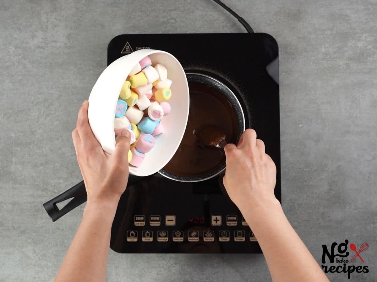 hand holding white bowl of marshmallows above saucepan of chocolate