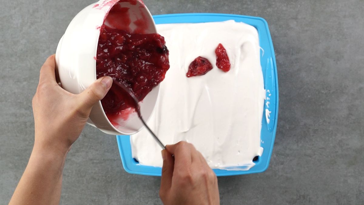 hand spooning cherry pie filling on top of whipped cream on cake