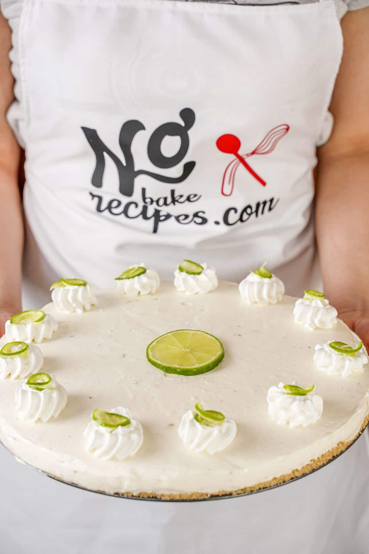 woman in apron that says no bake recipes holding whole key lime tart