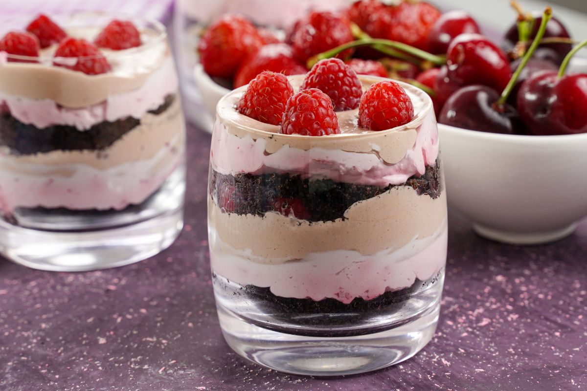 layered pudding parfaits with raspberries on top next to white bowl of fruit