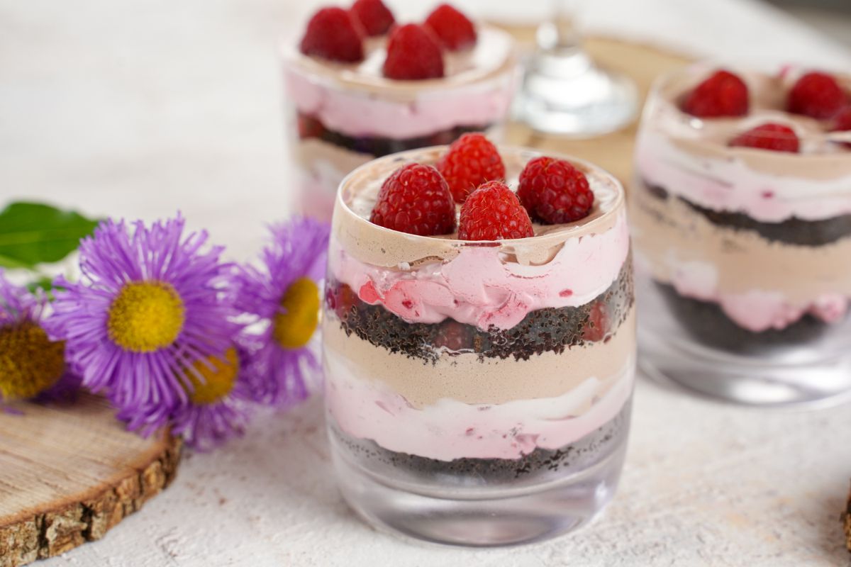 three glasses of Oreo pudding parfait topped by raspberries next to wood slice with purple flowers