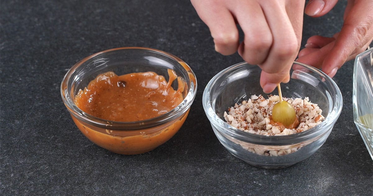grape on a toothpick with caramel on it being dipped into crushed nuts