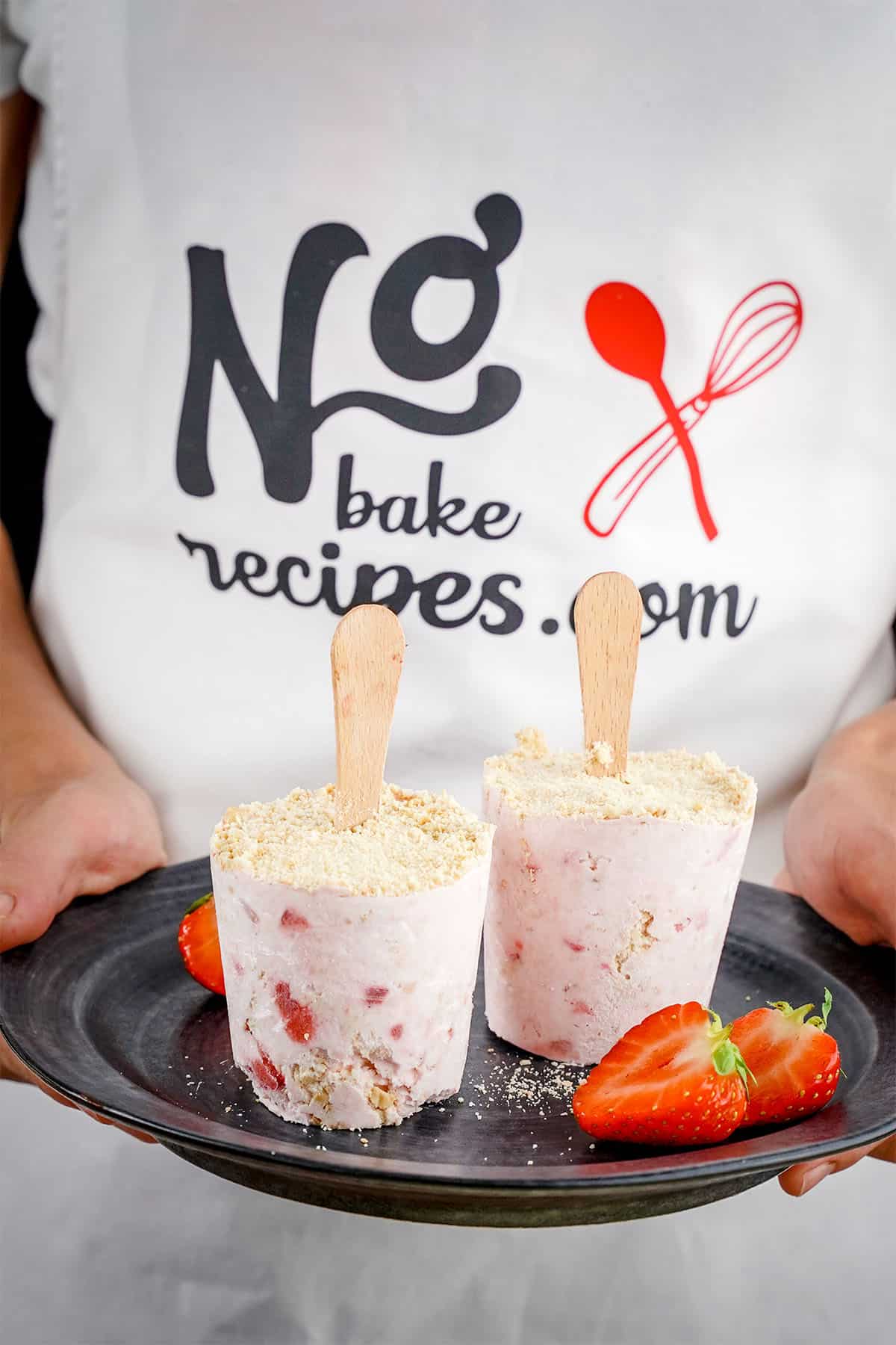 two strawberry cheesecake popsicles on a plate being held by someone wearing a no bake recipes apron